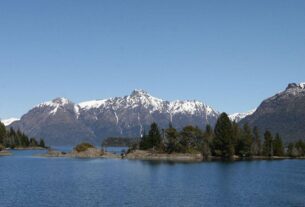 Mesa com câmera, caderno e xícara diante de uma janela com vista para lago e montanhas, em imagem ilustrativa de Bariloche.