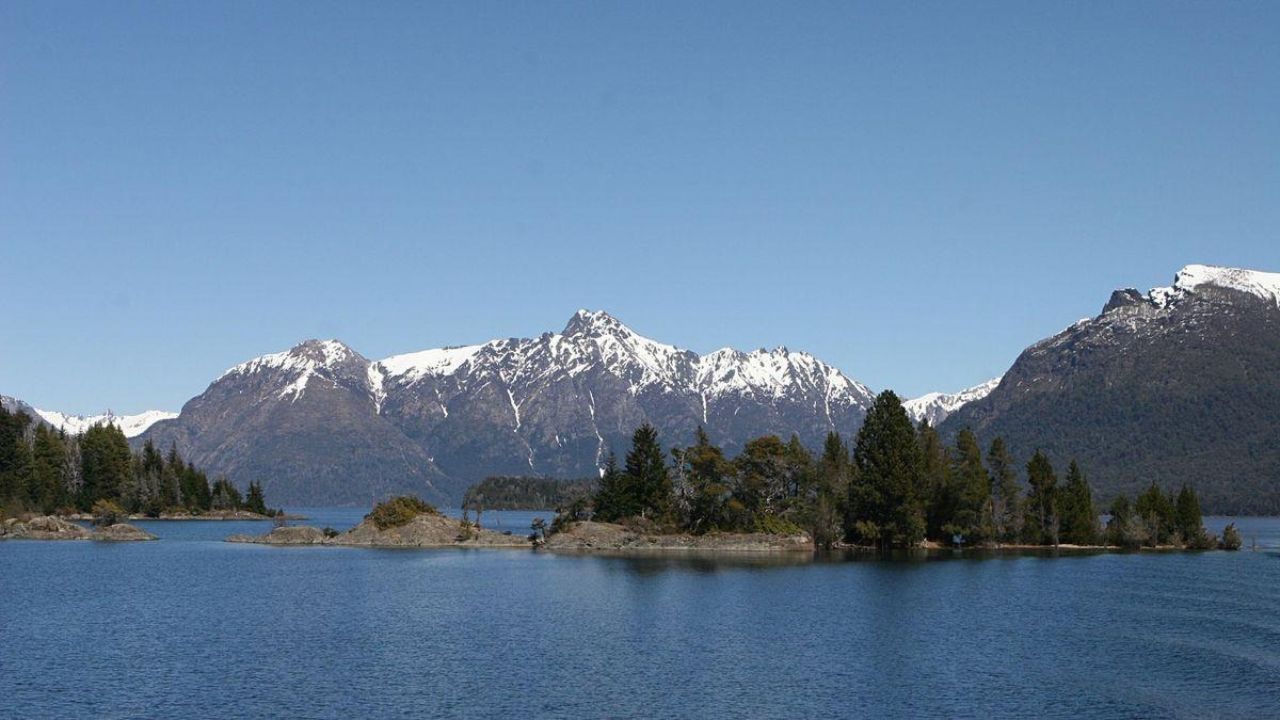 Mesa com câmera, caderno e xícara diante de uma janela com vista para lago e montanhas, em imagem ilustrativa de Bariloche.